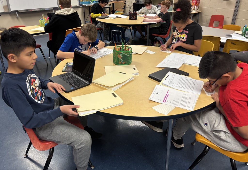 A group of students seated at round tables in a classroom, working on writing assignments and using laptops. Papers, folders, and writing tools are scattered across the tables as the students focus on their tasks.