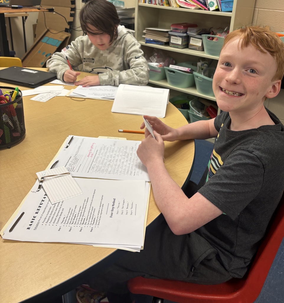 Two students are seated at a table working on writing assignments. One student smiles at the camera while holding an index card, and the other concentrates on writing. Their workspace is filled with folders, paper, and pencils.