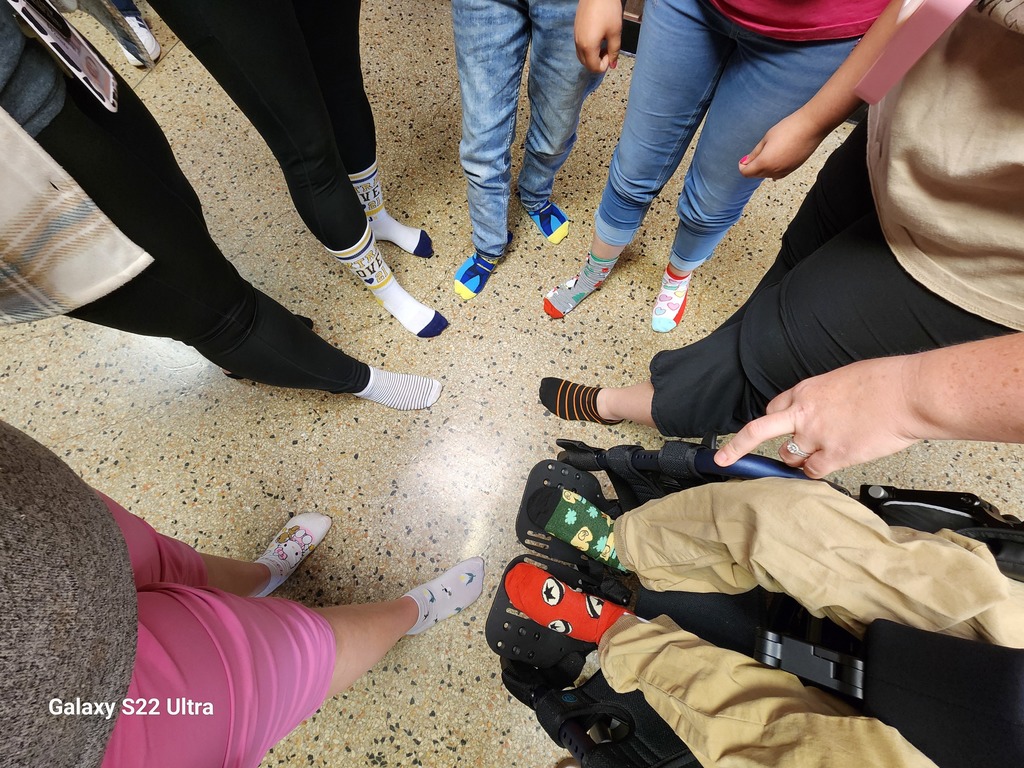 A close-up view of a group of people standing in a circle, showing off their mismatched and colorful socks on a speckled tile floor. The socks feature various patterns, colors, and characters. One person in a wheelchair has vibrant socks with cartoon faces. A hand is pointing towards them. The bottom left of the image contains the text 'Galaxy S22 Ultra.