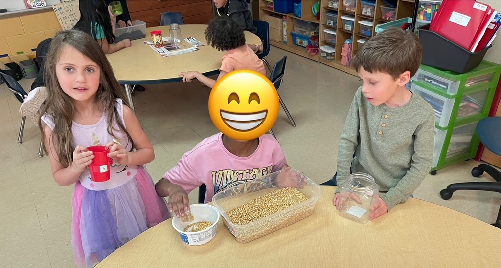 Three children at a classroom table work together measuring popcorn kernels with cups and plastic jars.