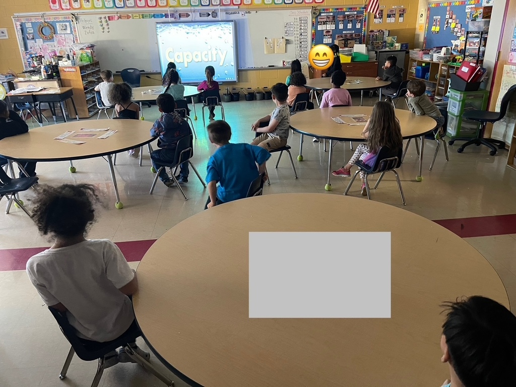 Children in a classroom sit at round tables and watch a screen at the front displaying the word “Capacity” in large text.