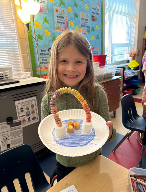 A girl in a green sweater happily holds up her completed cereal rainbow project on a decorated paper plate with marshmallows, cereal, and chocolates.