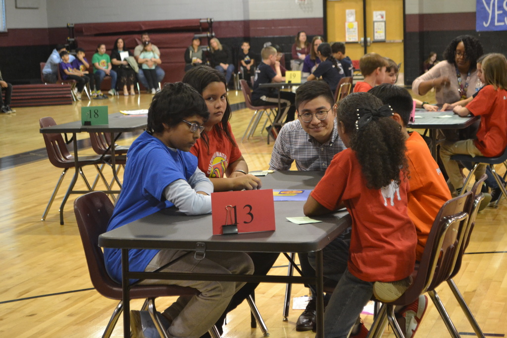 A group of students sits around a table marked with a red “13” sign during the Culpeper Math 24 Tournament. They are focused on the game in the center of the table, and a coach or teacher kneels beside them, smiling. Other teams and spectators are visible in the background of the gymnasium.