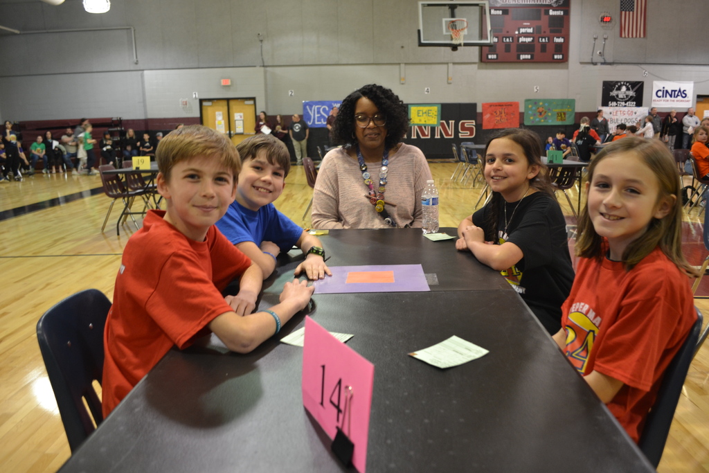  Four students sit around a table marked with a pink “14” sign during the Culpeper Math 24 Tournament. They are smiling and appear ready to play. An adult coach or judge sits at the head of the table, watching over the game. The gym is filled with other teams and colorful posters in the background.