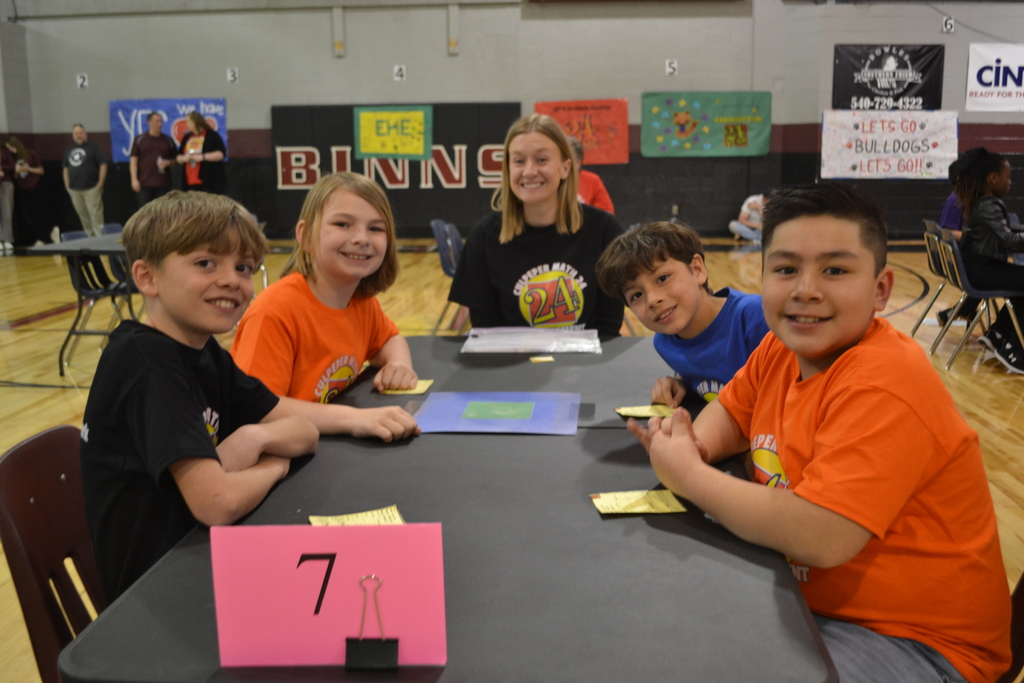 A group of five students, wearing Math 24 Tournament shirts in various colors, sit around a table labeled with a pink sign showing the number 7. They are smiling and ready to play the Math 24 game.