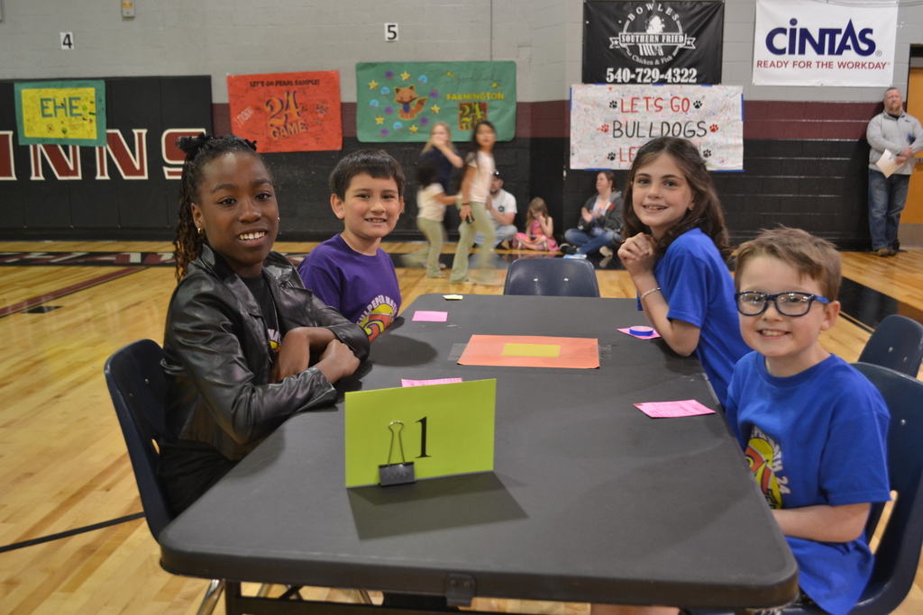 Four students wearing purple and blue tournament shirts sit at a table marked with a green number 1 sign. They are smiling at the camera, excited to compete.