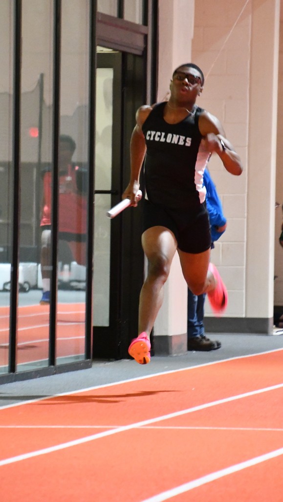 This is a photograph of a male athlete running an indoor track relay race. He is wearing a black sleeveless uniform with "CYCLONES" written in white across the chest, black running shorts, and bright pink track spikes. He holds a white relay baton in his right hand and appears to be in full stride, leaning forward with his left foot off the ground. He has a muscular build and is wearing black sunglasses. His facial expression shows focus and determination. In the background, there is a reflection of another person in the glass wall, and another runner wearing bright pink shoes is partially visible behind him. The indoor track has a bright orange surface with white lane markings.