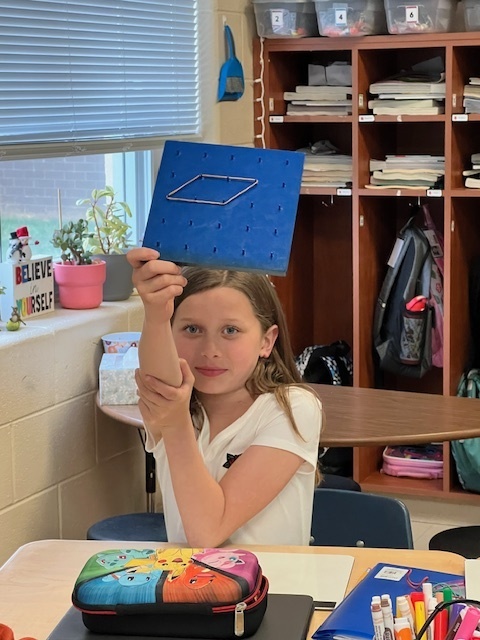 A girl proudly holds up a blue geoboard with a rubber band forming a rhombus shape. She is sitting at a desk with school supplies and student cubbies in the background.