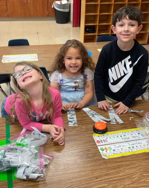  Three students standing at a classroom table, smiling at the camera while working on a hands-on activity involving strips of paper.