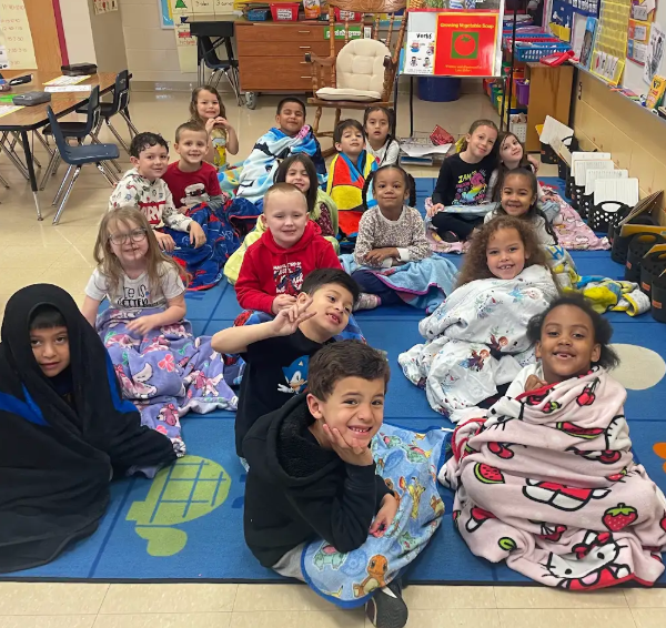 A classroom full of students sitting on a blue rug, many wrapped in colorful character blankets, smiling and posing for a group picture.