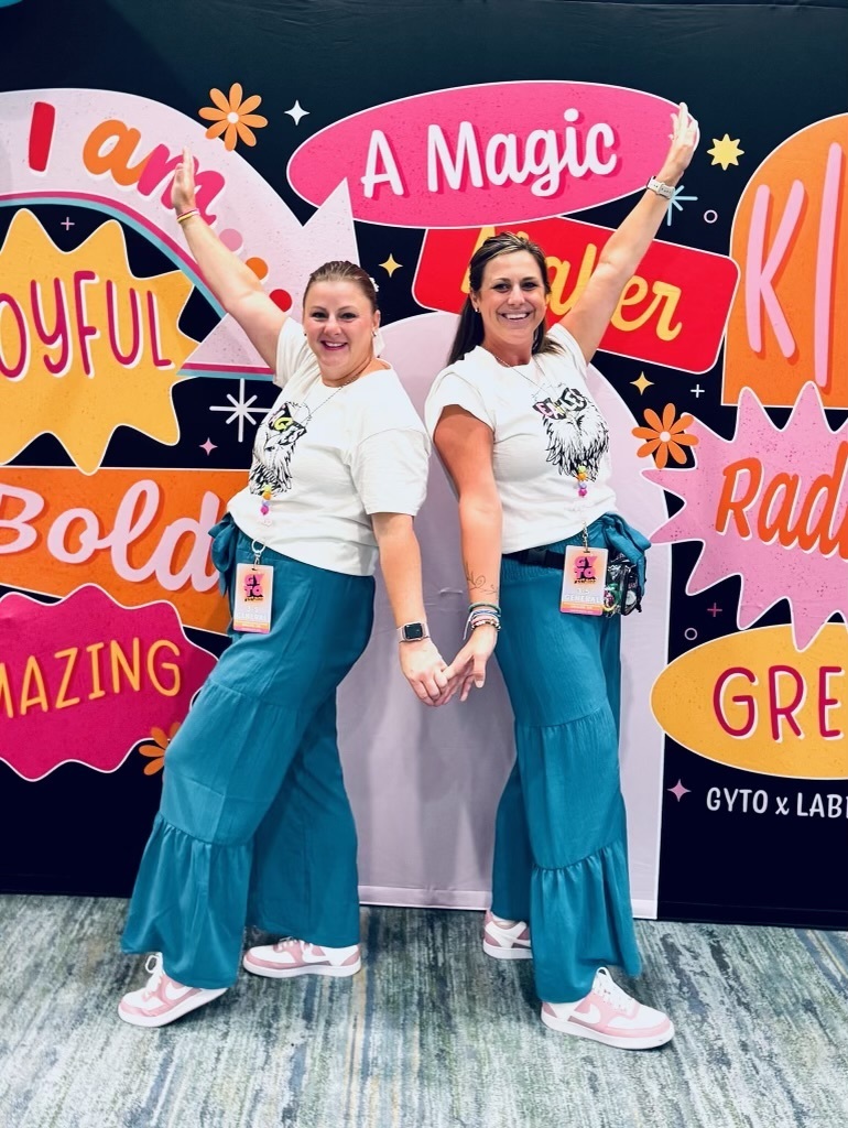 Two women stand back-to-back smiling and holding hands in front of a colorful backdrop with affirmations like "I am joyful," "A magic maker," and "Amazing." They are wearing matching outfits: white graphic T-shirts with a wildcat design, teal tiered pants, and pink-and-white sneakers. Each wears a lanyard with a name tag, and both have one arm raised enthusiastically. The backdrop includes logos for GYTO x LAB.