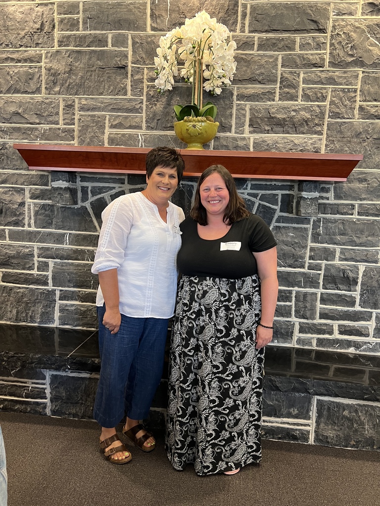 Two women stand side by side smiling in front of a stone wall with a dark wooden mantel. A yellow-green vase with white orchids sits on the mantel above them. 