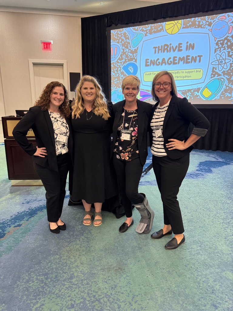 Four women stand together smiling in a conference room in front of a screen displaying a presentation titled “Thrive in Engagement: Guide for Assistant Principals to Support and Encourage Participation.” All four are dressed in professional attire, with three wearing blazers and the fourth in a black dress. One woman wears a medical walking boot on her right leg. The background features a projector, podium, and colorful carpeted floor.