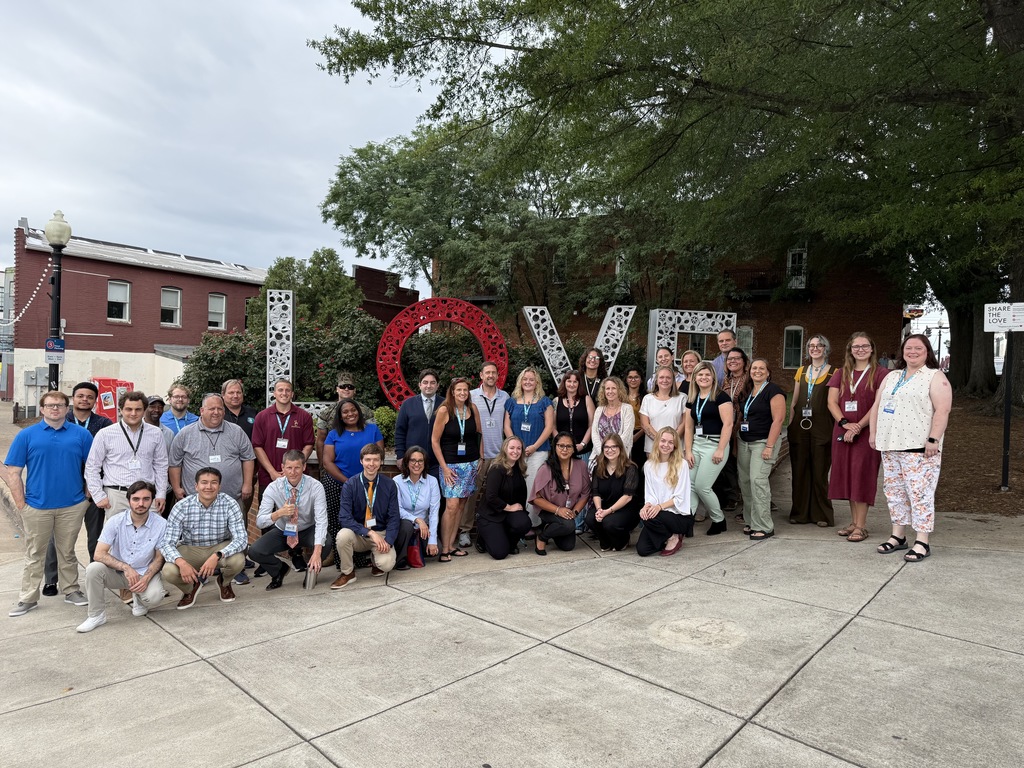 A large group of new Culpeper County Public Schools teachers pose together in front of the "LOVE" sculpture in Downtown Culpeper. The group is smiling and standing on a sidewalk surrounded by trees and brick buildings,