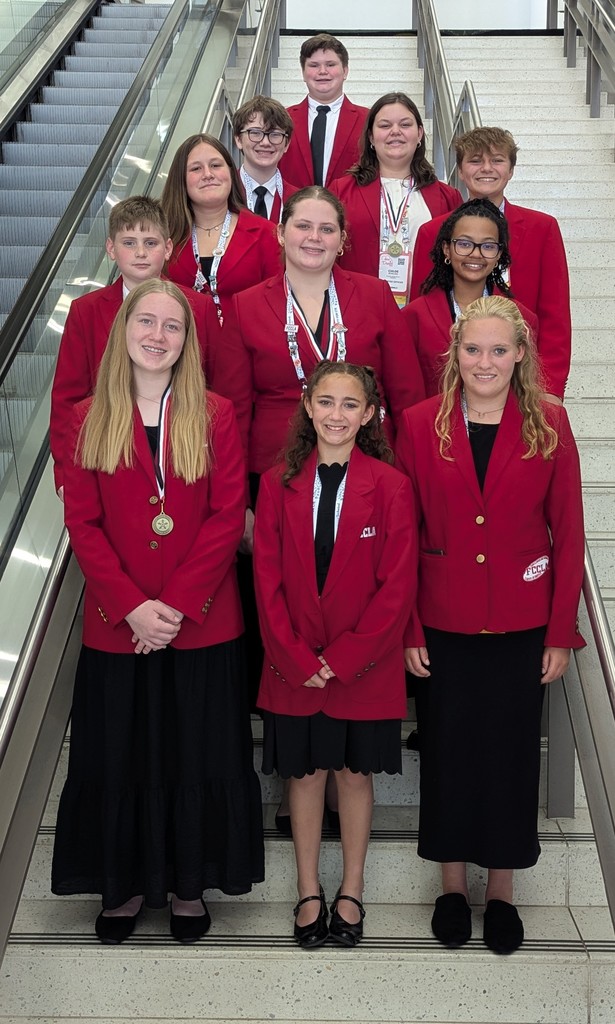 A group of 13 students wearing matching red FCCLA blazers poses on a staircase between two escalators. The group consists of boys and girls of various ages, all dressed in formal attire with black bottoms. Several students wear medals and lanyards, indicating participation or achievement in an event. They are smiling and standing in organized rows, representing their organization with pride.