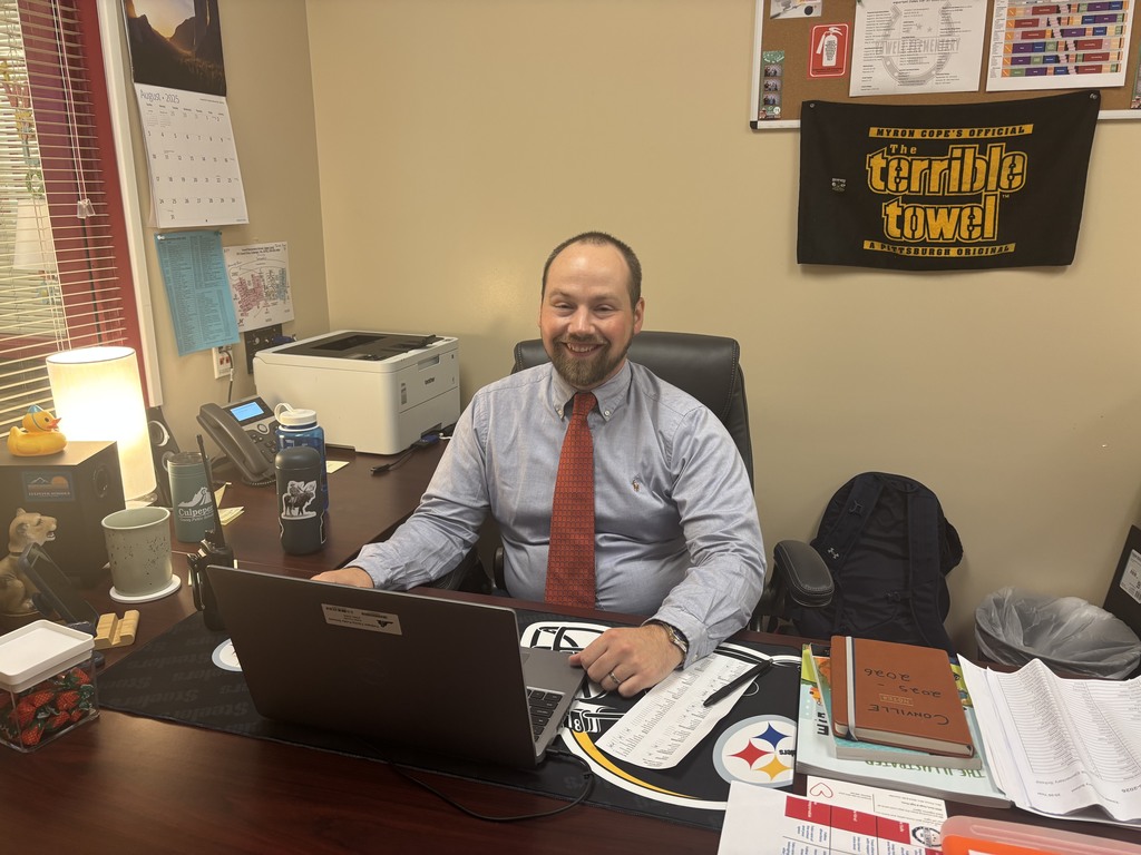 Mr. Conville, Yowell Elementary’s new dean of students, sits smiling at his desk in an office. He is wearing a light blue dress shirt and a red tie, and is seated in front of a laptop. His desk has various personal items, including a Steelers mousepad, a water bottle with a wolf sticker, a walkie-talkie, mugs, a rubber duck, and a container of wrapped strawberry candies. Behind him on the wall are a bulletin board with schedules and a "Terrible Towel" banner, as well as a calendar and printed maps.