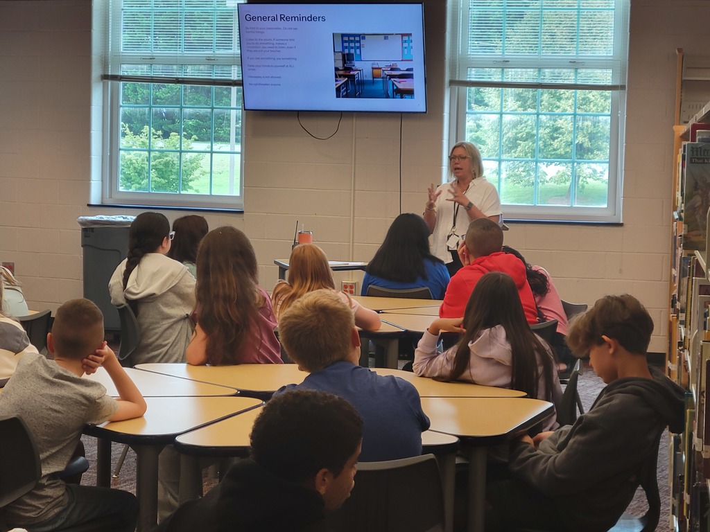 A teacher stands at the front of a classroom, speaking to a group of middle school students seated at grouped tables. The students are attentively listening as a slide titled "General Reminders" is displayed on a large screen behind the teacher. The room has large windows that let in natural light, and there are bookshelves and posters visible along the side.