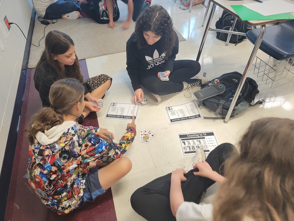 A group of five middle school students sit in a circle on the classroom floor, engaged in a card game. They are using printed activity sheets titled "Counting to 14" and "Use Doubles to Make 10 or 20." One student wears a colorful anime-themed hoodie. Nearby are backpacks, notebooks, and a desk. The classroom has a tiled floor with a maroon border and white walls.