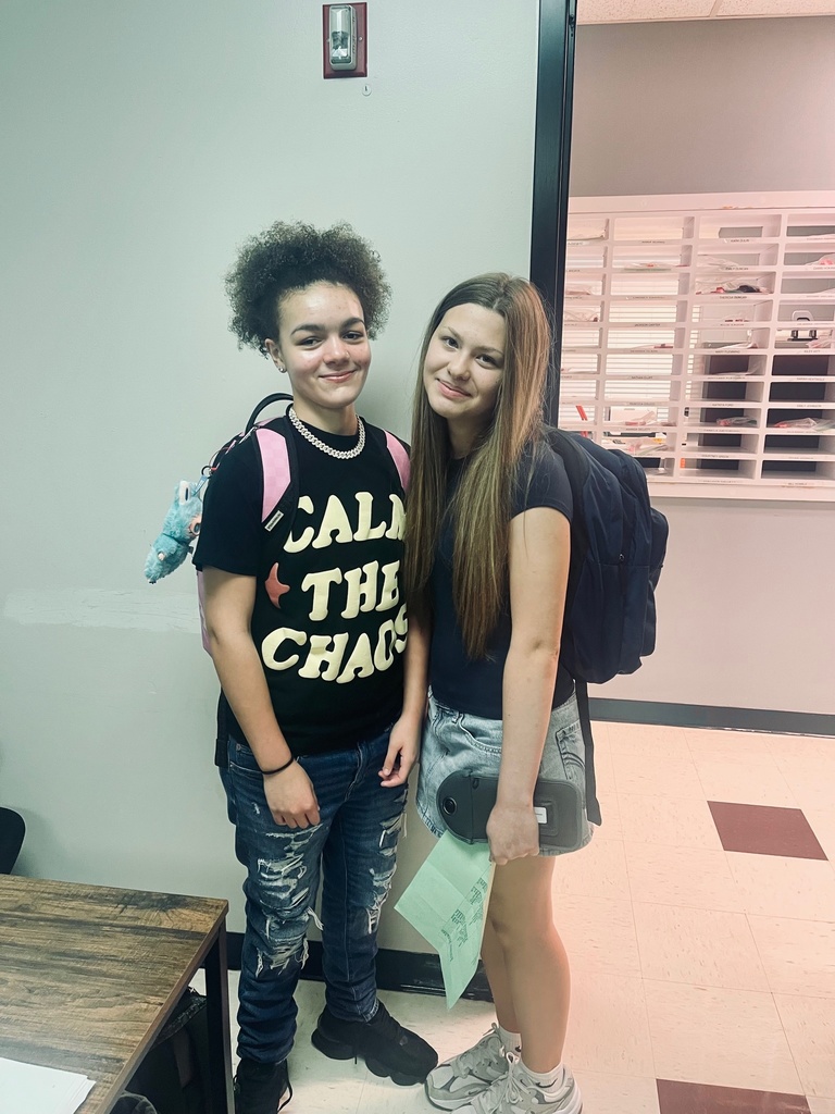 Two middle school students stand and smile for a photo in a school hallway. One student wears a black t-shirt that says "CALM THE CHAOS" and ripped jeans, while the other wears a black t-shirt and a denim skirt, holding a green paper and a pencil pouch. Both have backpacks on and are standing near a classroom door with cubby-style mail slots visible in the background.