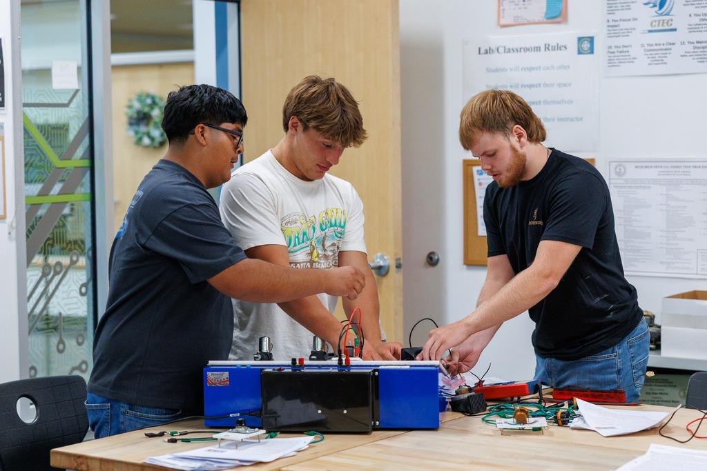 Three students collaborate on an electronics project in a CTEC classroom, working with wires and components connected to a blue circuit board device on a wooden workbench. They appear focused and engaged, surrounded by tools, papers, and lab equipment. Lab/classroom rules are posted on the wall behind them.
