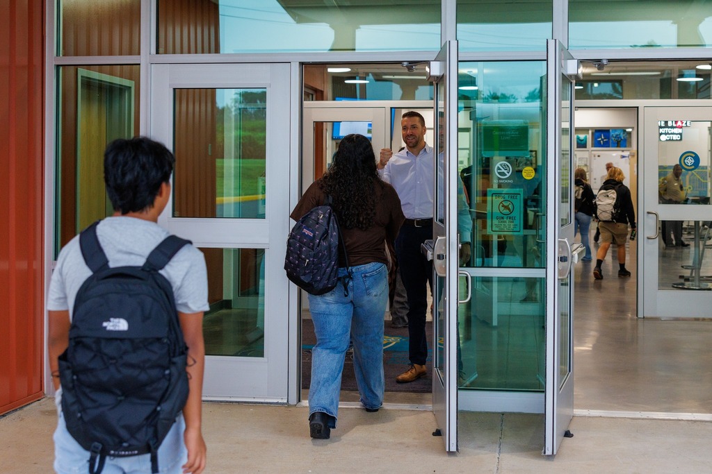 A staff member stands at the entrance of CTEC, smiling and raising a fist in a welcoming gesture as students arrive on the first day of school. One student with a patterned backpack walks through the open glass doors, while another student with a black backpack follows closely behind. More students are visible inside the building, walking toward their classrooms. 