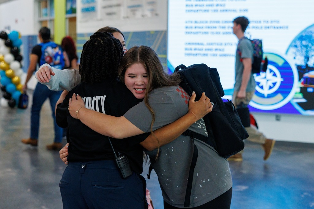A staff member enthusiastically greets students as they walk into the main entrance of CTEC on the first day of school. One student, wearing a brown shirt and blue jeans with a backpack, approaches the door while another student follows behind. More students are visible entering the building in the background.
