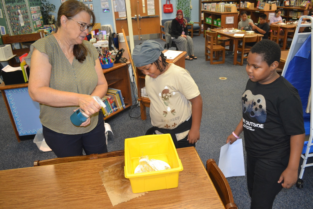 Image contains the adult spraying a project designed by two students as they watch in the school library.  Other students working in the background while and adult is sitting in a chair supervising.