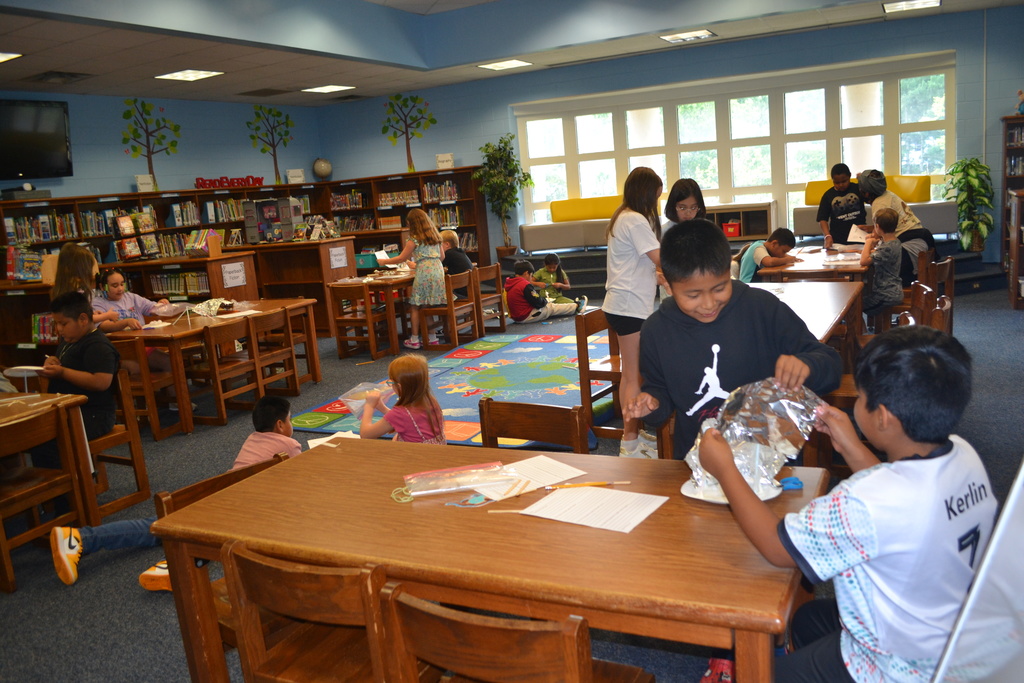A class of students in the library working together on a STEAM project.  Students are spread out around different tables with their supplies.
