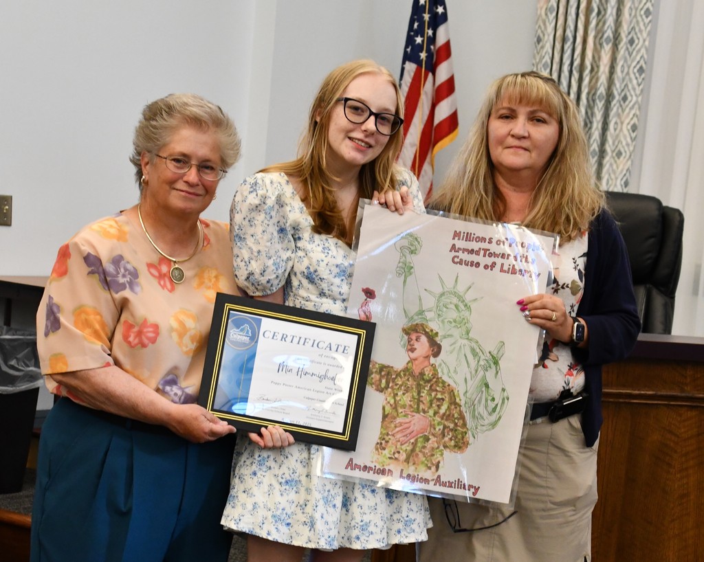Mia Himmighoefer, Culpeper County High School graduate, stands holding a certificate and her award-winning Poppy Poster from the American Legion Art Contest, alongside a School Board member and teacher Marla Bell during a school board meeting.