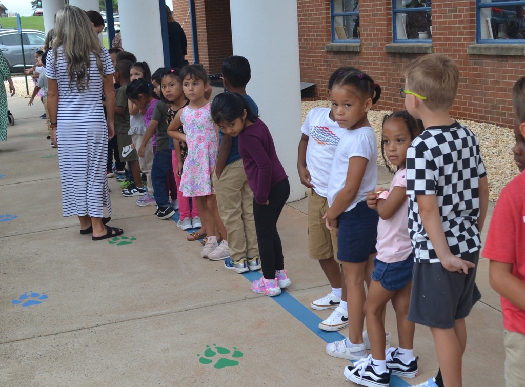 A group of students practice waiting on the blue painted line in front of the school during a bus safety drill.  A teacher and other staff are seen in the background.