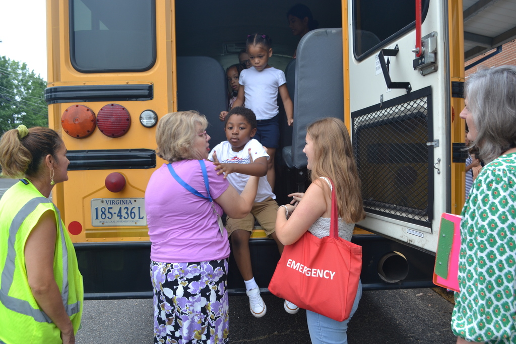 A bus driver and teacher watch as other staff help a student safely learn how to evacuate during a bus drill.