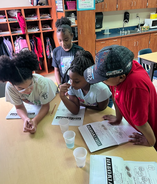 Four students gather around a classroom table, looking at worksheets titled “Experiment.” They observe cups of liquid on the table while discussing. The classroom background includes cubbies, a sink, and cabinets.