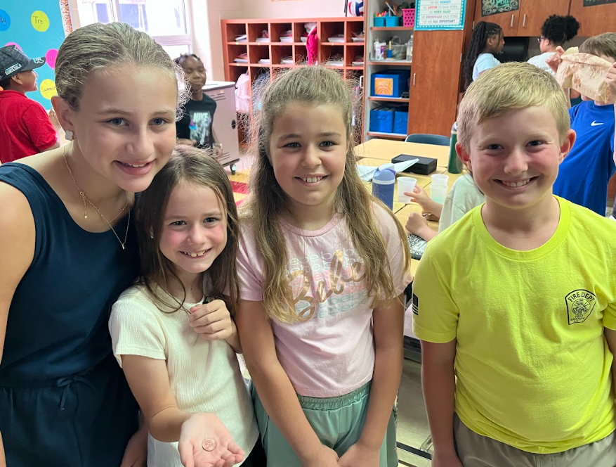 Four smiling students stand together in a classroom, posing for the camera. One student in the front holds a small object in her open palm. Other students and colorful classroom decorations are visible in the background.