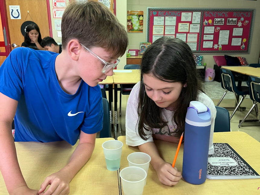 Two students work together at a classroom table, closely examining cups of liquid. The boy in a blue shirt leans over while the girl with long dark hair writes with a pencil. A water bottle and notebook are on the table.
