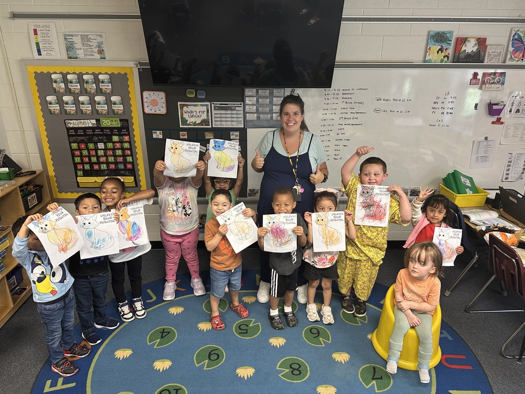 Teacher poses with a group of preschoolers in their classroom on the rug.  Each student is holding up their colored fox proudly showing off their work.