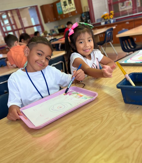 A boy and girl sit side by side at a classroom table, smiling while holding paintbrushes. Both have papers with colorful circular designs in front of them.