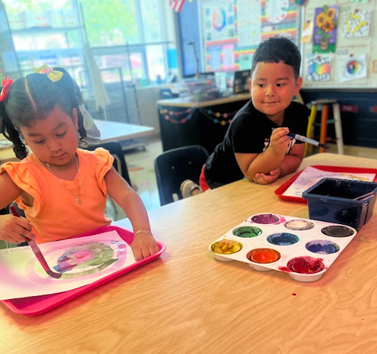 Two children sit at a classroom table with watercolor palettes. The girl in an orange shirt paints on her paper while the boy in a black shirt looks on with his brush in hand.