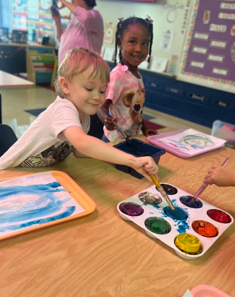 Two children sit at a classroom table, one boy painting with a brush dipped in blue while a smiling girl in a tie-dye shirt works beside him. A watercolor palette with bright colors is in front of them.