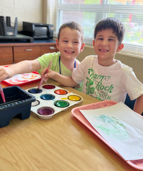 Two smiling boys sit together at a classroom table, holding paintbrushes over a watercolor tray. One wears a light green shirt, and the other wears a white shirt with green writing. A sheet of painted paper rests on the table.