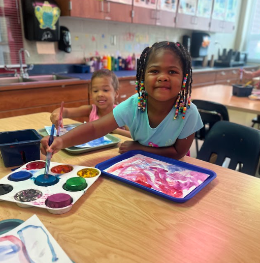 Two smiling girls sit together at a table, painting with bright watercolors. The girl in front, with colorful beads in her hair, looks at the camera while using blue paint.