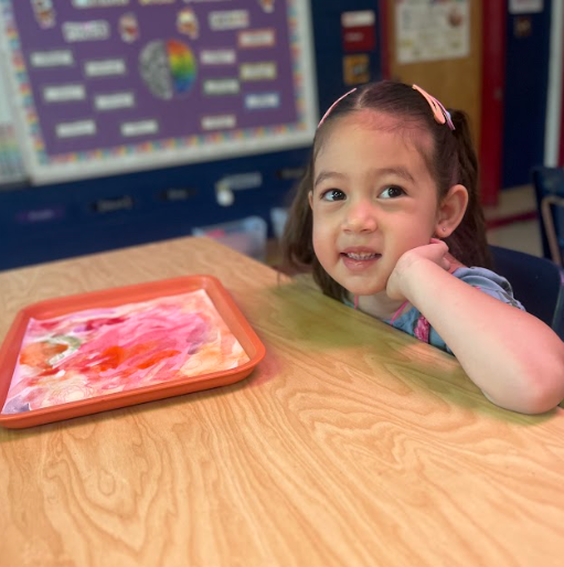 A young girl leans her chin on her hand and smiles at the camera, sitting at a classroom table with a tray holding a bright red and pink watercolor painting.