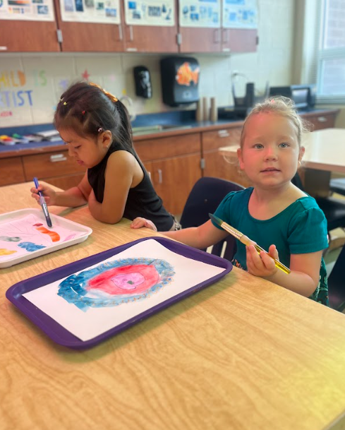 Two girls sit side by side at a classroom table, painting with brushes. The girl in front, wearing a teal shirt, looks at the camera, and her paper shows a circular watercolor design.