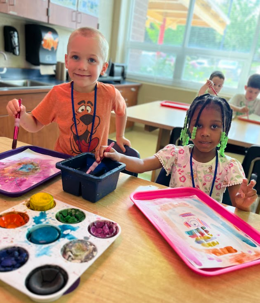 Two young children sit at a classroom table using paintbrushes and trays of watercolors. The boy wears an orange Scooby-Doo shirt and smiles, while the girl with green beads in her hair holds up her hand and looks at the camera.
