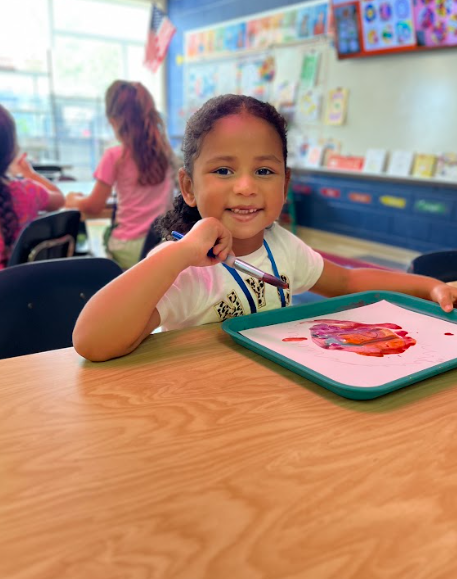 A young girl sits at a classroom table smiling at the camera, holding a paintbrush over a tray with a colorful watercolor painting in shades of red and purple.