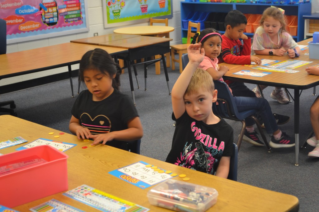 Image contains 5 young students sitting at their tables.  Boy student has his hand raised.  Students are focused and working on a bingo game.