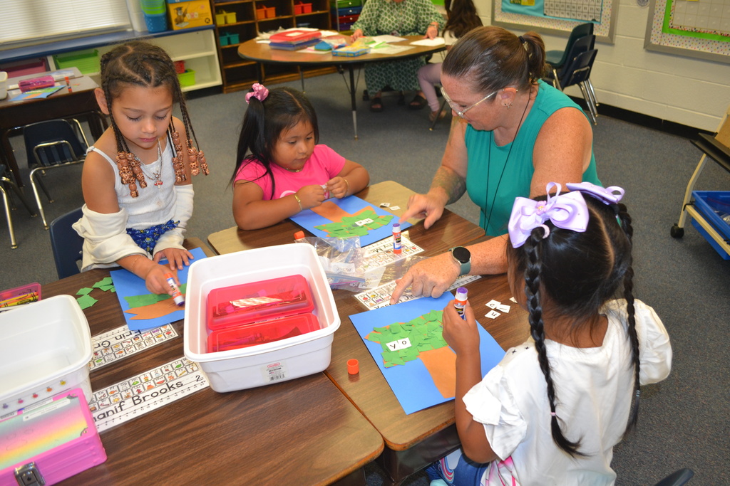 Image contains a teacher working with three students while they are a their desks.  Students are creating a craft with construction paper and glue sticks.