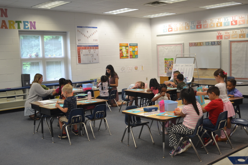Image contains a classroom with tables.  Students are seated in their chairs and working.  3 adults are bent down at 3 tables helping to assist the students.