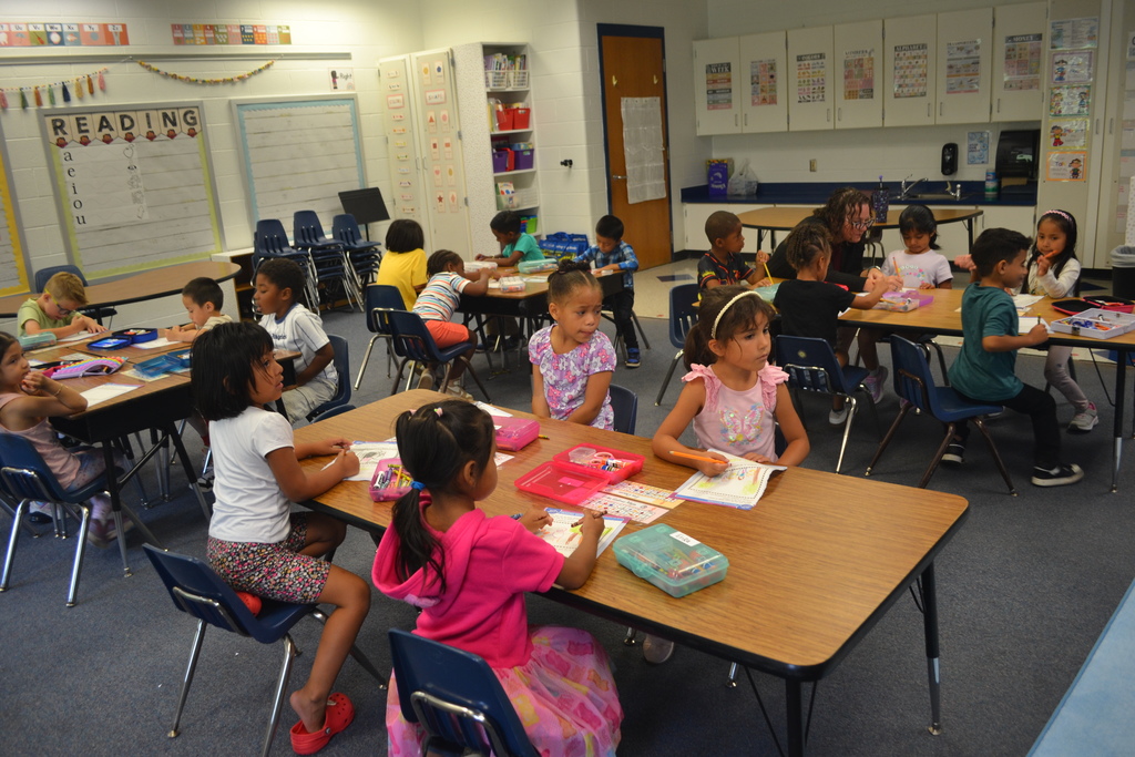 Image contains a classroom full of children.  Each child is sitting at their spot and are working and coloring an activity.  1 adults is pictured helping a student.