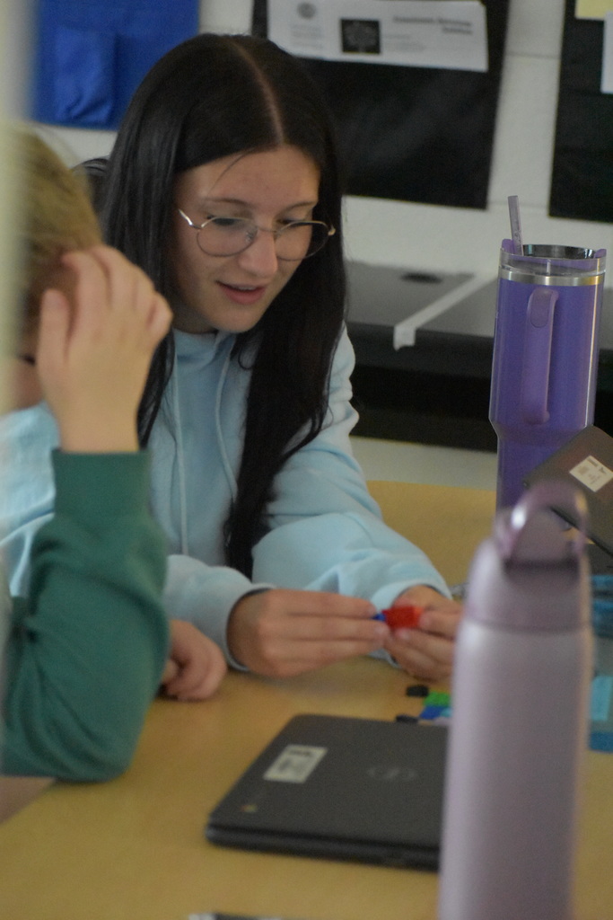 A student with long dark hair and glasses, wearing a light blue hoodie, is seated at a desk and holding small red and blue plastic pieces. Another student in a green sweatshirt is partially visible beside them. Two large reusable water bottles and a closed Chromebook are on the desk. The background shows classroom materials and papers on the wall.