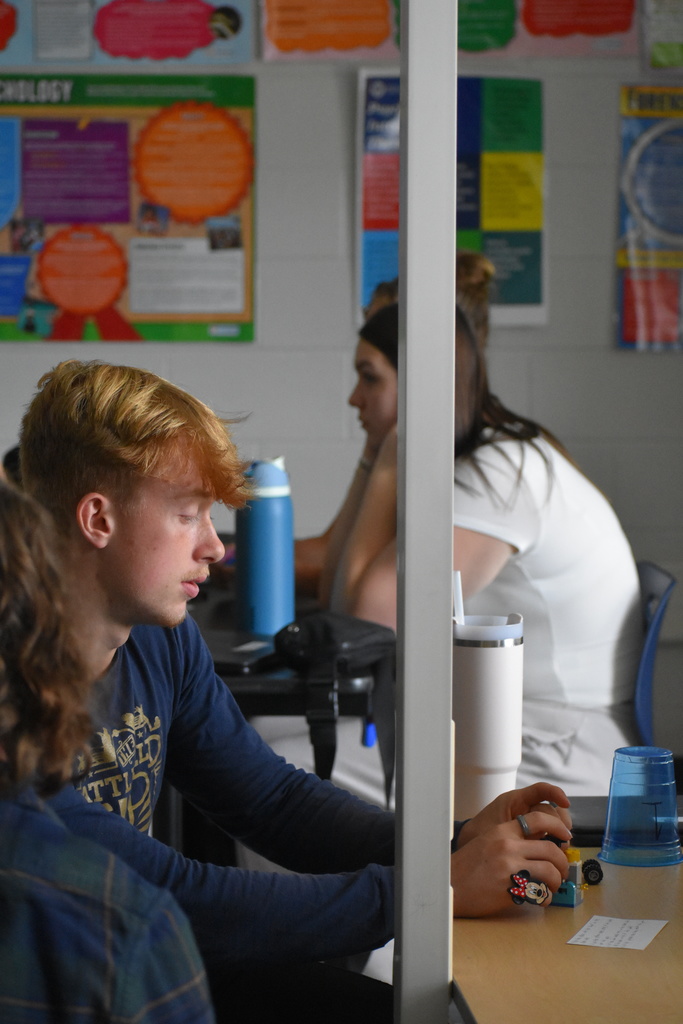 A student with light brown hair wearing a navy long-sleeve shirt sits at a desk, focused on small Lego pieces. A white tumbler and a blue plastic cup are on the desk, and colorful posters hang on the wall in the background.